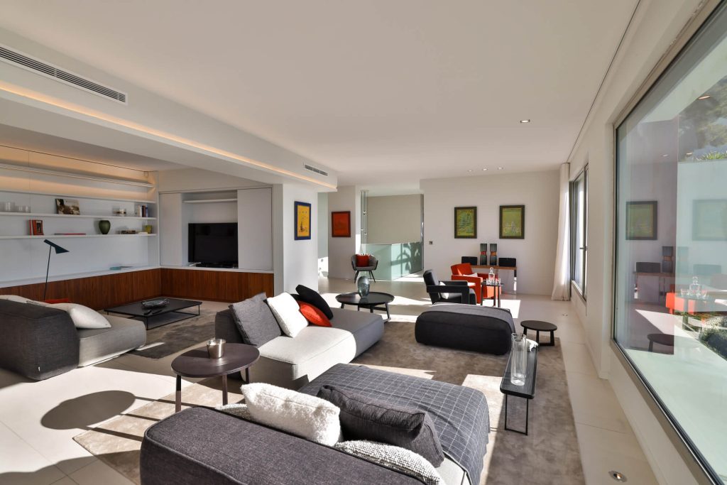 Bright, modern living room with contemporary decor, large picture windows, gray and beige designer sofas, colorful cushions, minimalist furnishings, recessed lighting, desk area in background with orange chairs, framed works of art, sleek, elegant ambience, upscale interior with outdoor views.