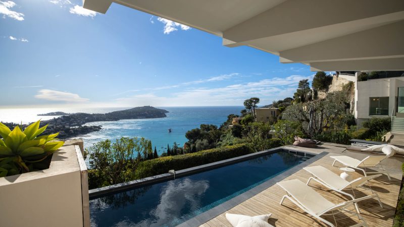 Terrasse de villa de luxe sur la Côte d’Azur avec piscine à débordement, transats design, vue panoramique sur la mer Méditerranée et la presqu’île de Saint-Jean-Cap-Ferrat, ciel bleu ensoleillé et cadre verdoyant.