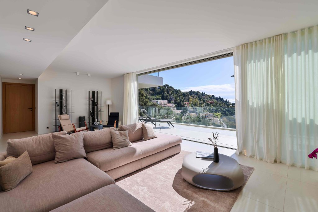 Bright, modern living room in a luxury villa on the Côte d'Azur with panoramic views of the green hills, large bay window, beige designer sofa, contemporary furniture, sunny terrace and minimalist decor.