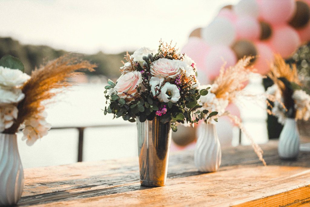 Floral centerpiece of pastel roses, eucalyptus and dried flowers on a rustic wooden table. A refined setting for an outdoor wedding by the water. 