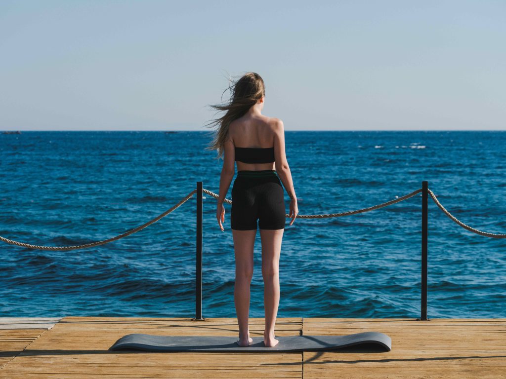 Young woman in sportswear facing the sea, standing on a yoga mat on a wooden deck. Open-air wellness session, between fitness and meditation, at the water's edge. 