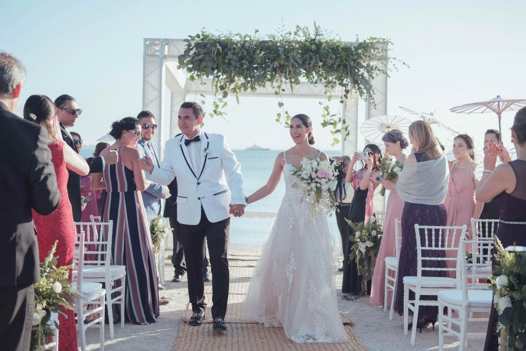 A radiant bride and groom walk hand in hand under a seaside flower arch, surrounded by their guests. Elegant, romantic wedding ceremony on the beach. 