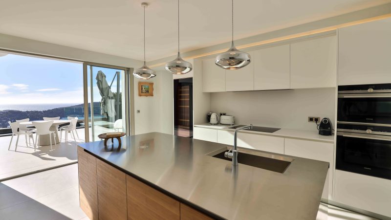 Grande cuisine moderne avec îlot central en bois et plan de travail en inox, éclairée par trois suspensions chromées. Une baie vitrée s’ouvre sur une terrasse avec table blanche et vue panoramique sur la mer